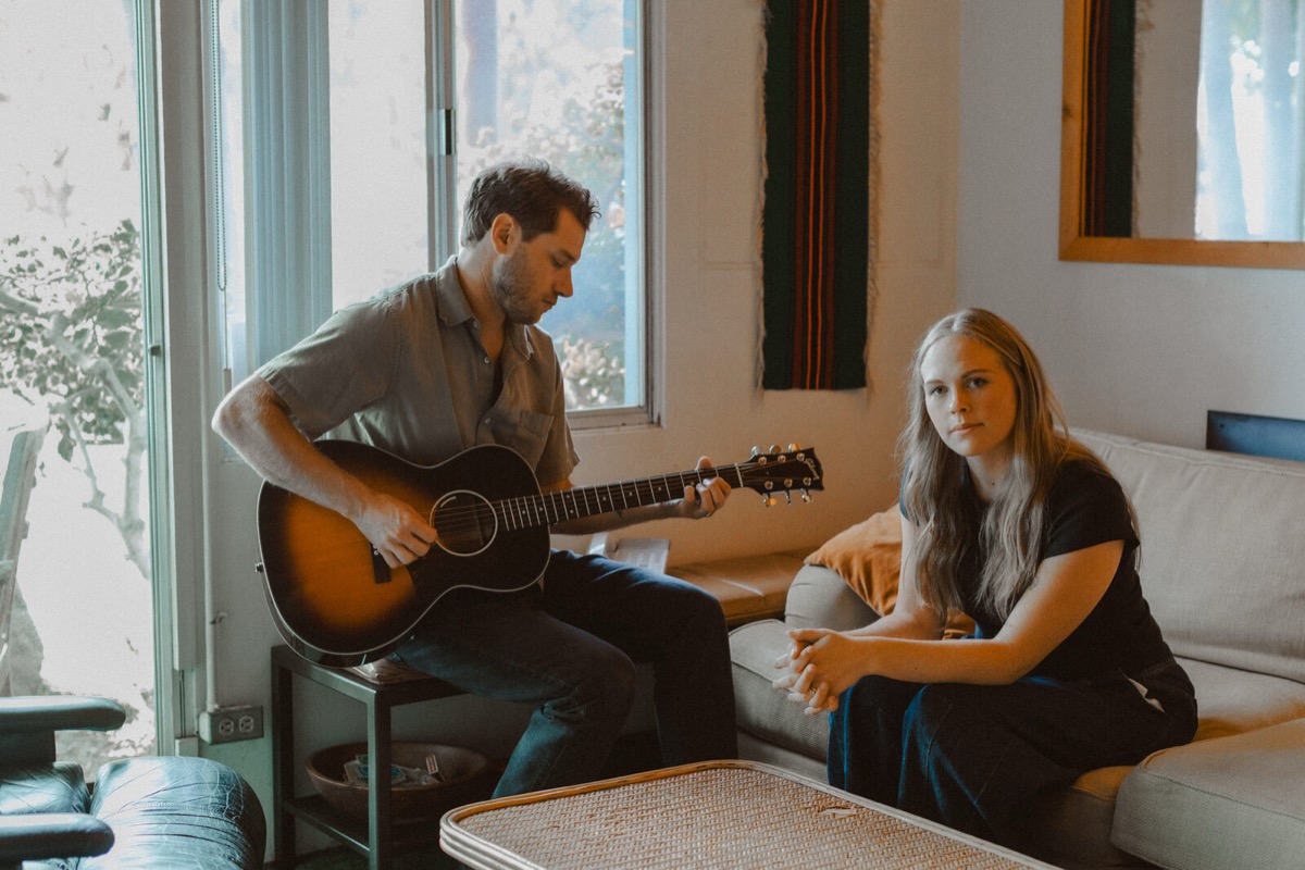 Brian playing acoustic guitar beside Kadi in a sunlit living room.